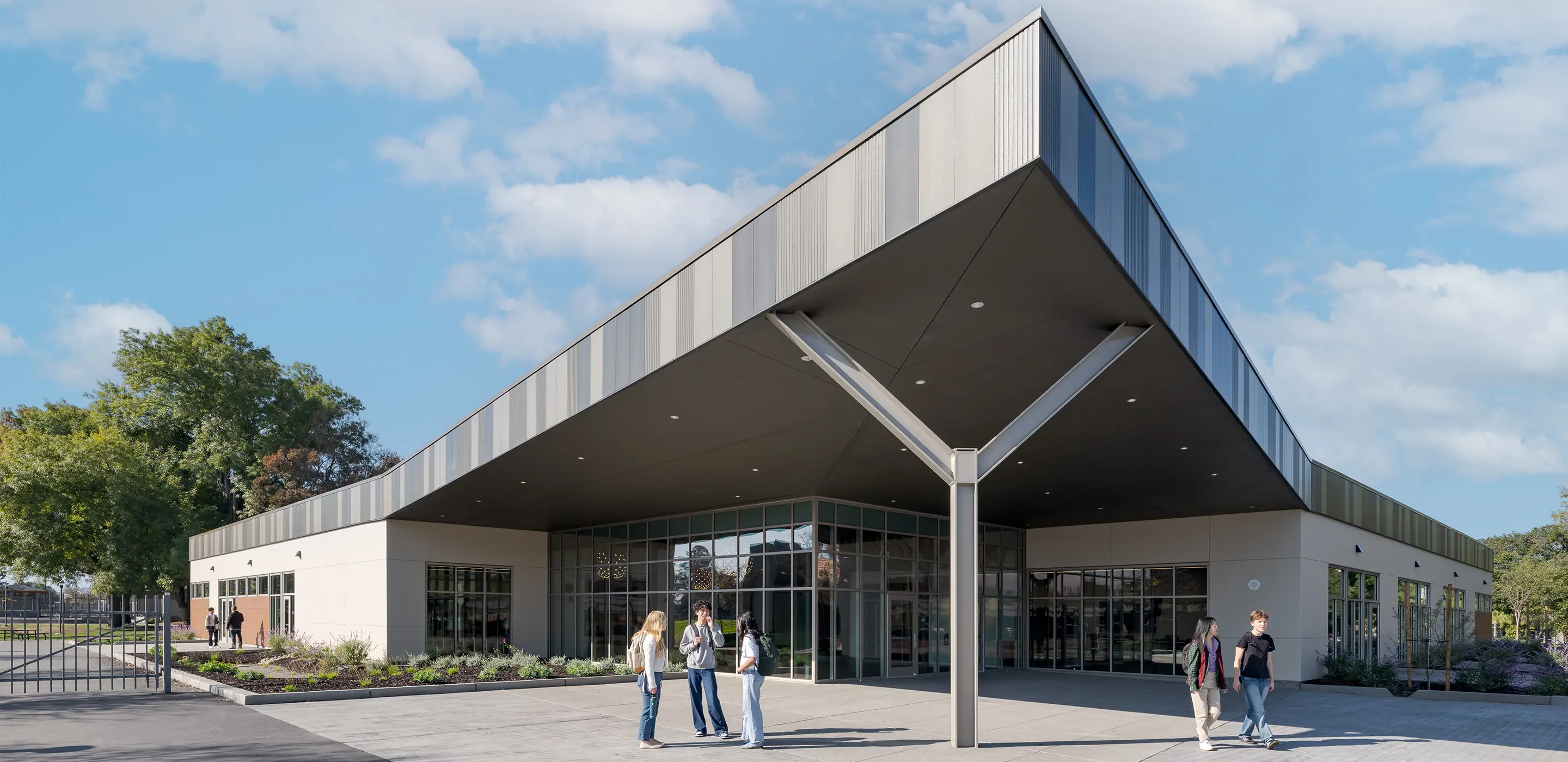 Modern building with angular roof, glass facade, and students walking and talking outside under a blue sky.