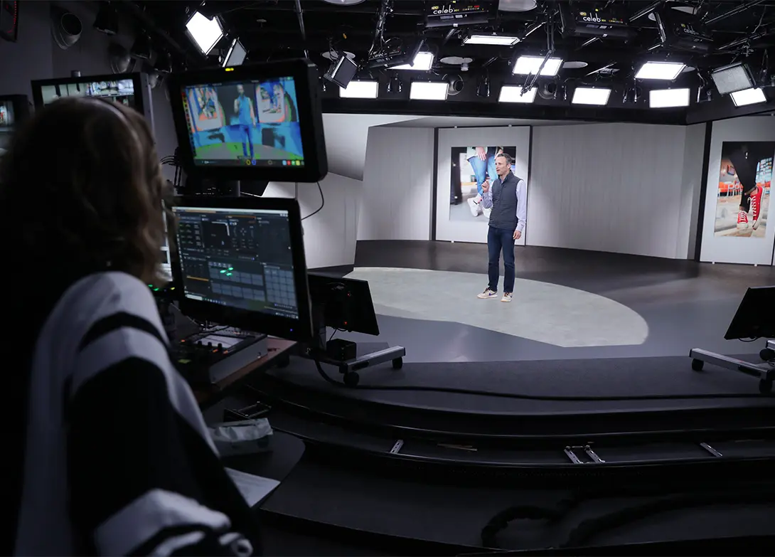 Person presenting in a professional studio, with operator at control panel, multiple screens and lights visible.