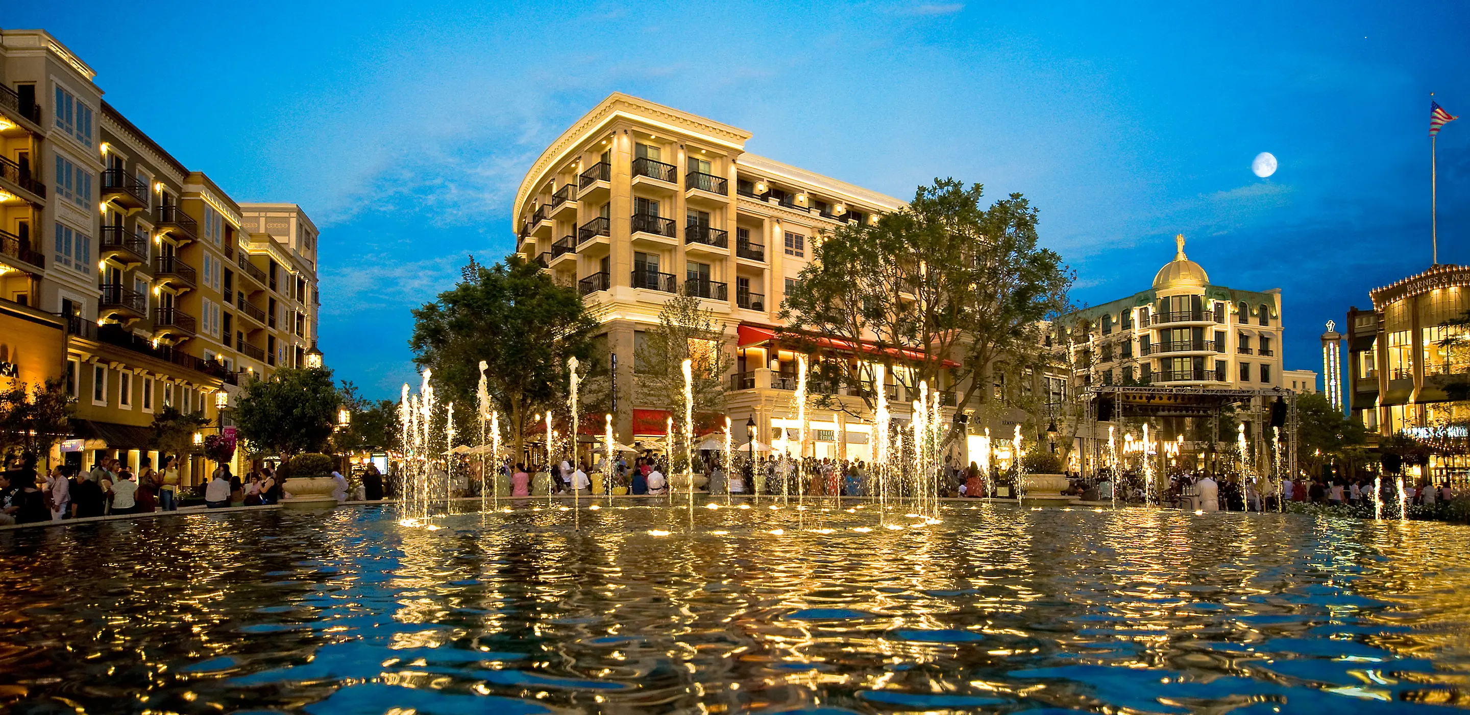 Outdoor fountain and illuminated buildings at night, reflecting in water, with a clear sky and moon in the background.