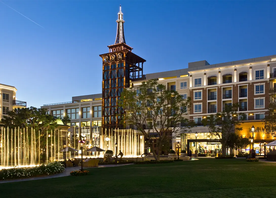 Night view of a shopping center with illuminated fountain and tower, surrounded by modern architecture and trees.