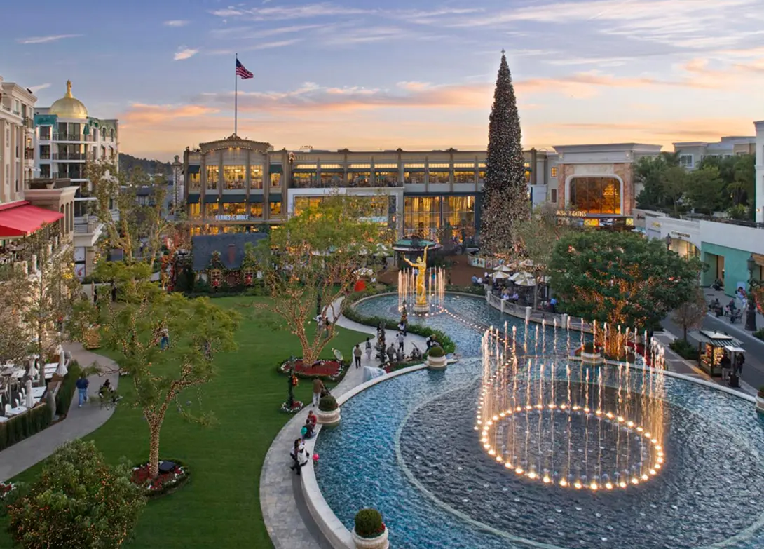 Outdoor shopping center with fountain and Christmas tree at sunset, featuring festive decorations and lush greenery.
