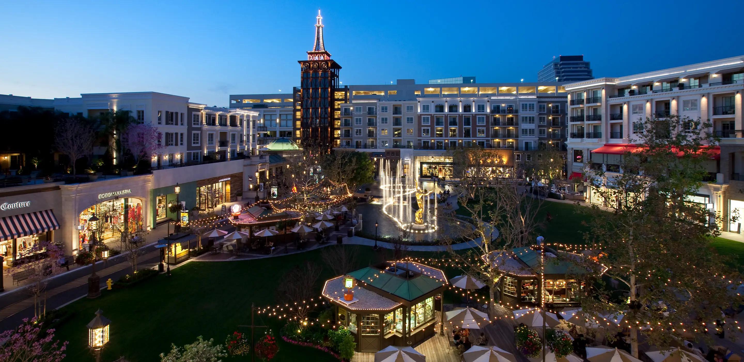 Vibrant evening view of an illuminated outdoor shopping mall with fountain and shops.