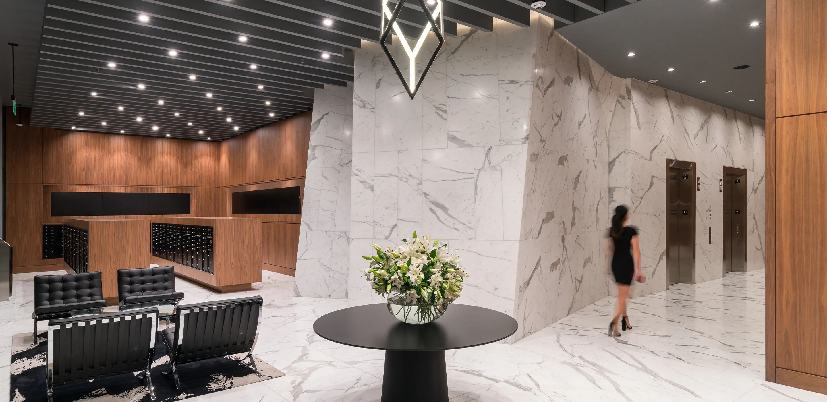 Modern office lobby with marble walls, wooden accents, seating, and flower arrangement. Woman walking to elevators.