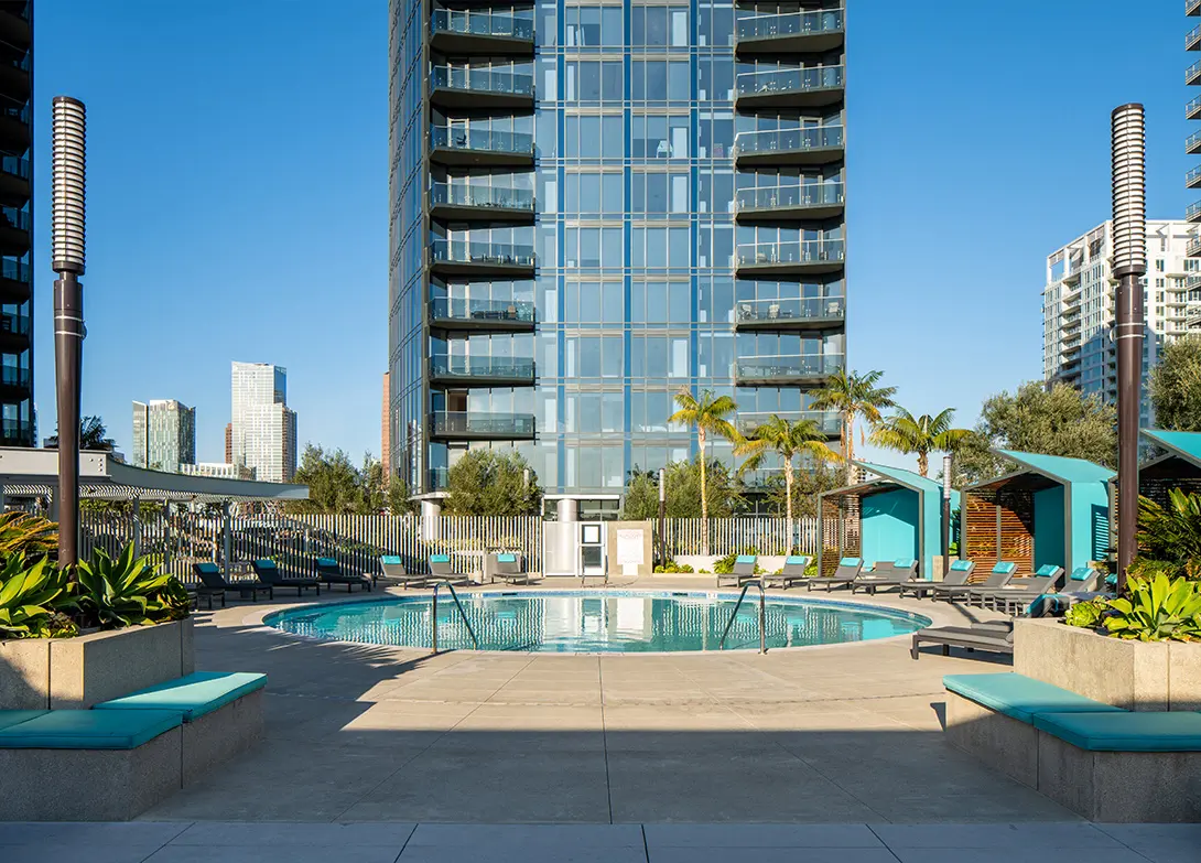 Modern apartment building with outdoor pool, lounge chairs, and palm trees under a clear blue sky.