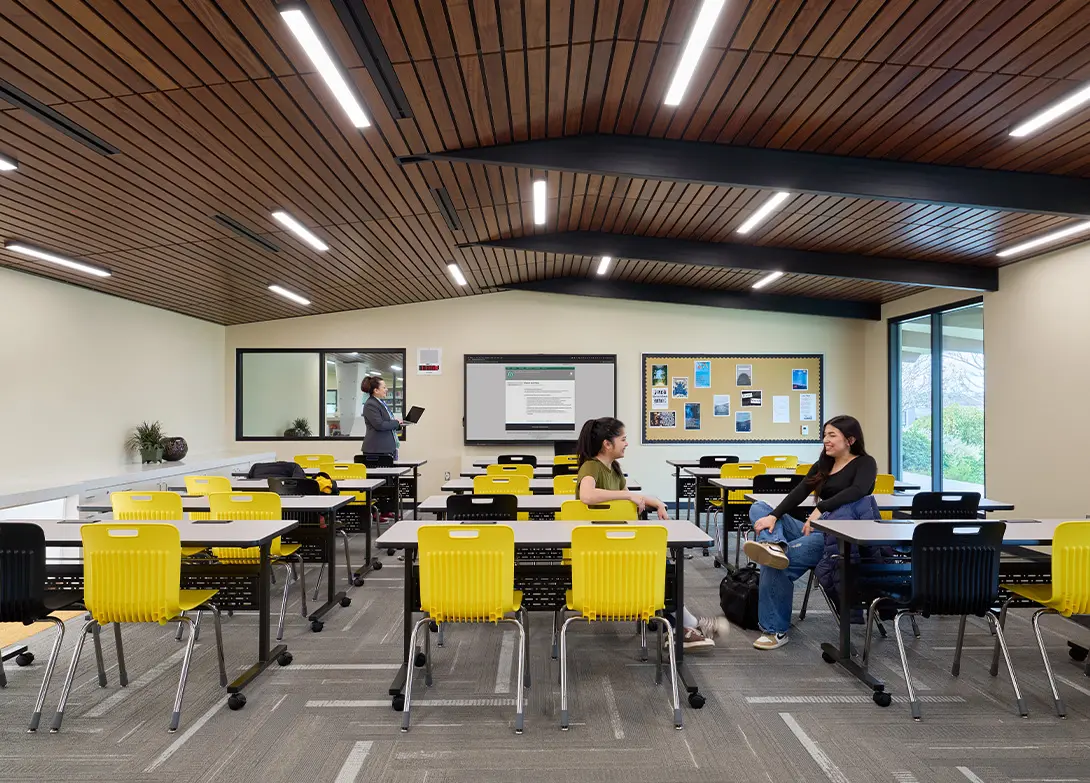 Modern classroom with yellow chairs, two students talking, and a person using a laptop near a smartboard.