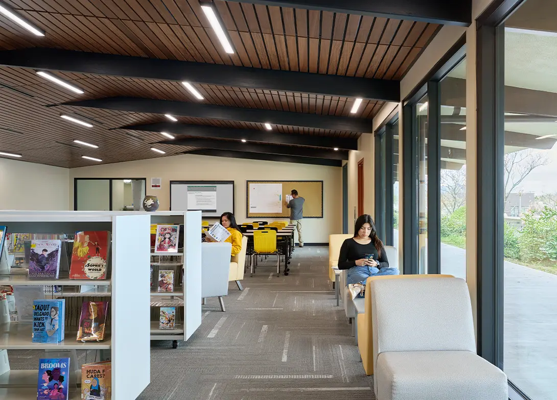 Modern library reading area with people studying and browsing books, featuring large windows and comfortable seating.