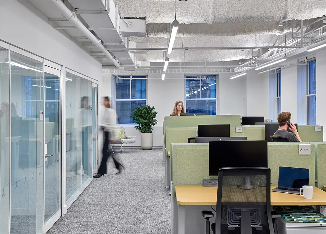 Modern office workspace with people working at cubicles, large windows, and glass-walled meeting rooms.