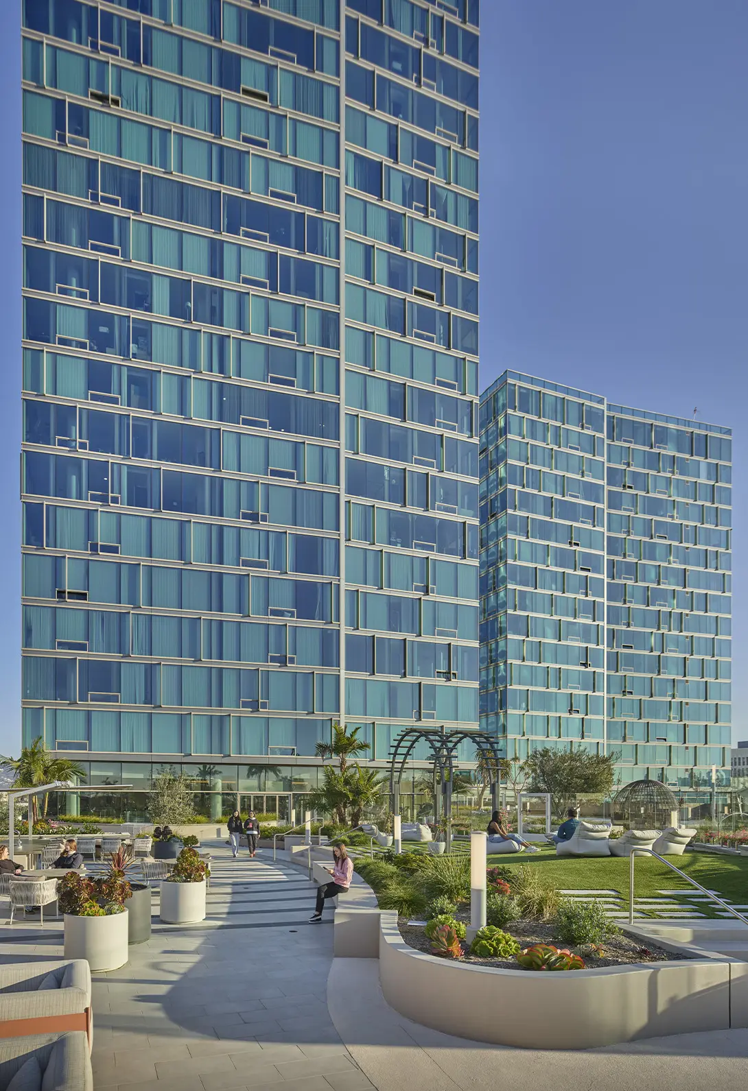 Modern urban skyscrapers with glass facades, surrounded by a landscaped garden and seating area under a clear blue sky.