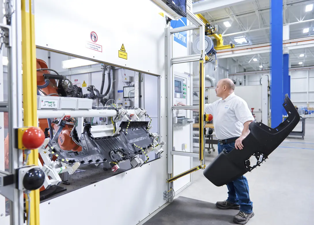 Factory worker operates robotic arm in manufacturing plant, holding car bumper.
