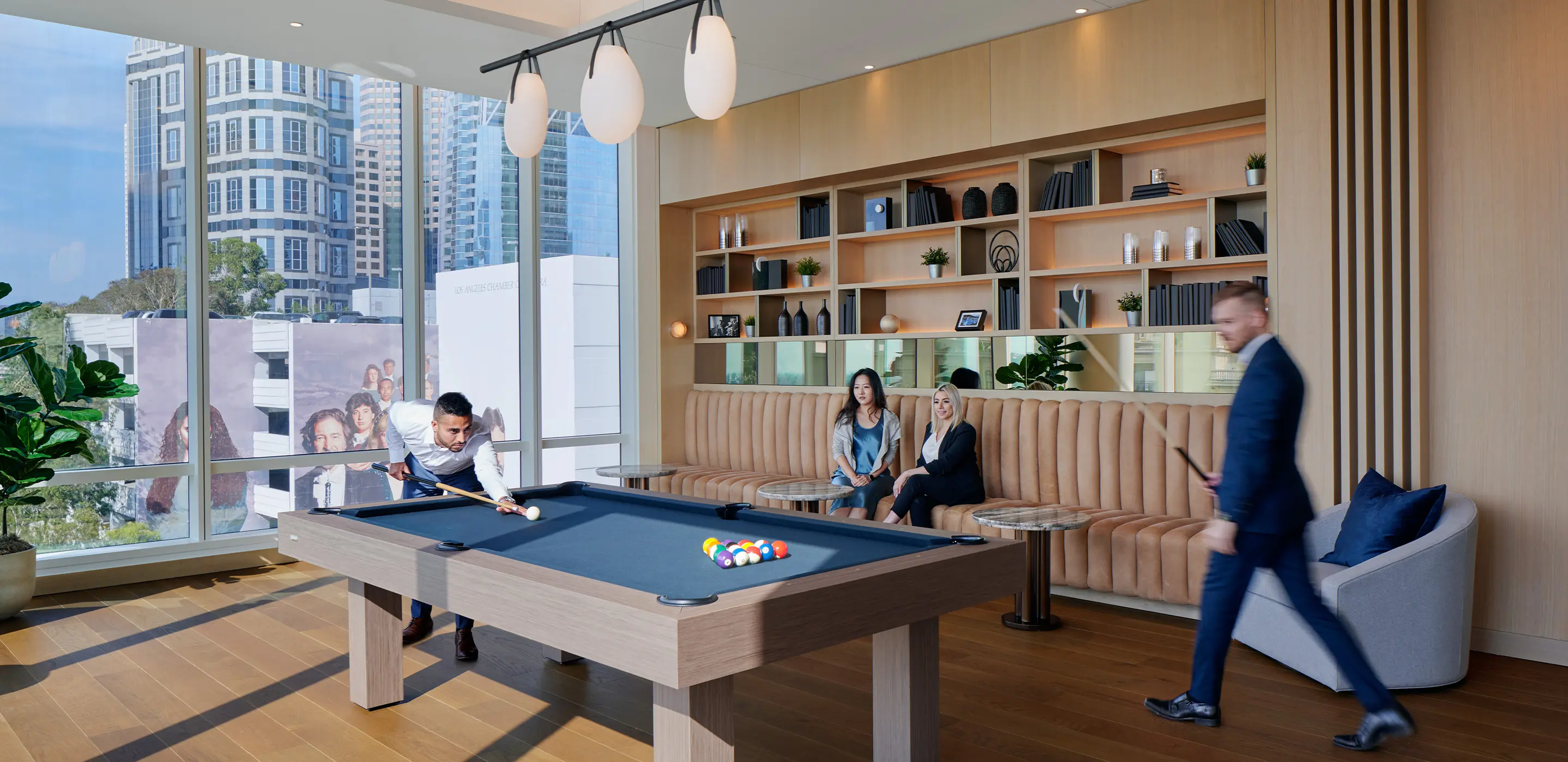 Modern lounge with people playing pool, city view windows, stylish seating, and wooden flooring.
