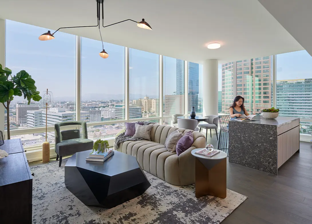 Modern living room with large windows, city views, stylish furniture, and a person reading at the kitchen island.