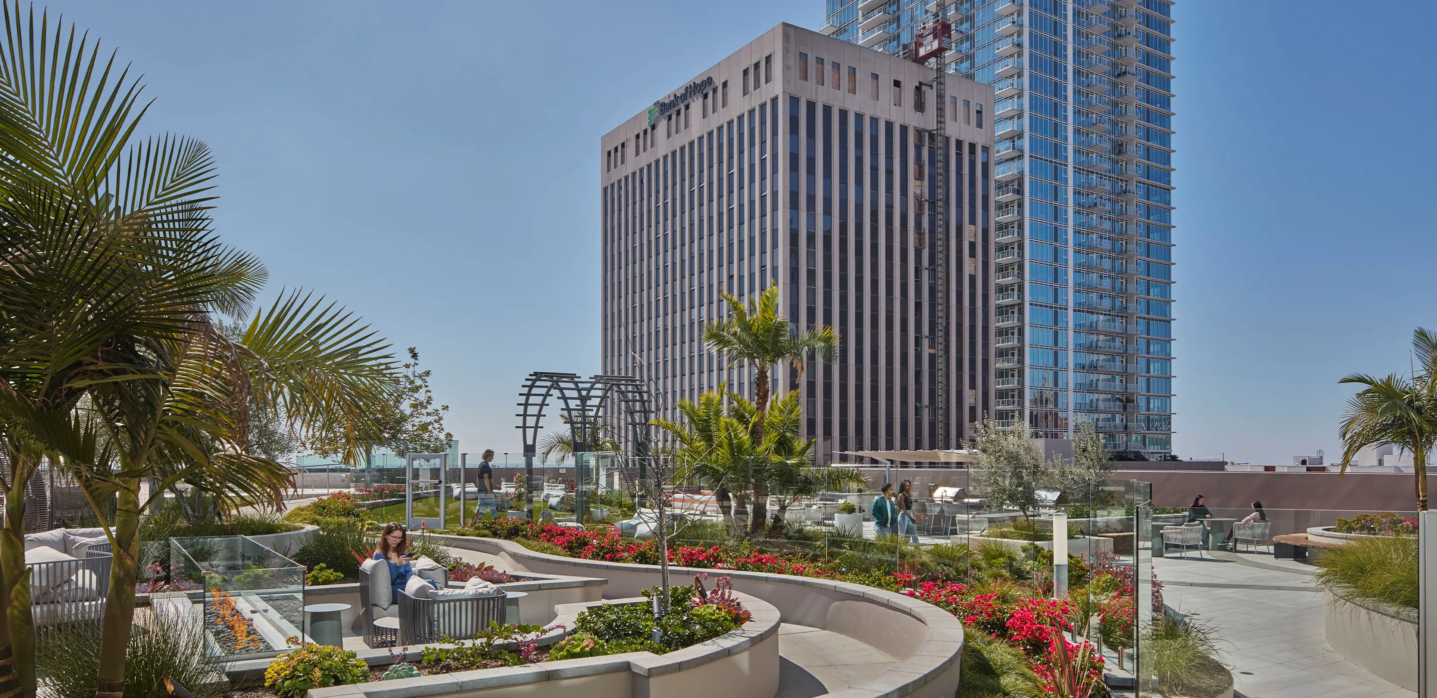 Rooftop garden with seating, palm trees, and flowers, overlooking city skyscrapers under a clear blue sky.