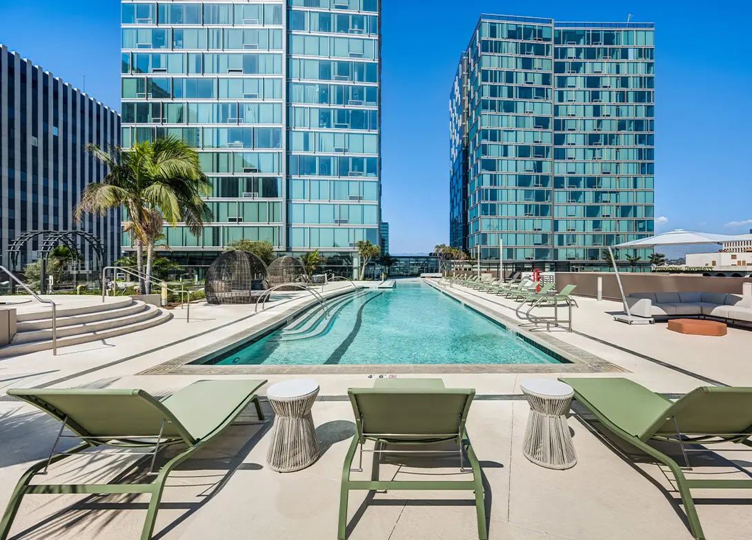 Luxury apartment pool area with lounge chairs, modern high-rise buildings, and palm trees under a clear blue sky.