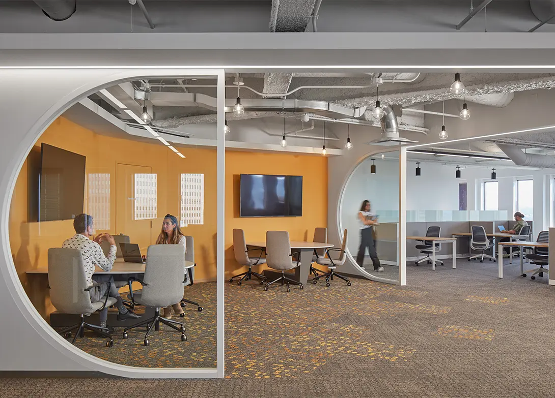 Modern open-plan office with meeting spaces, vibrant orange accent wall, and circular glass partitions.