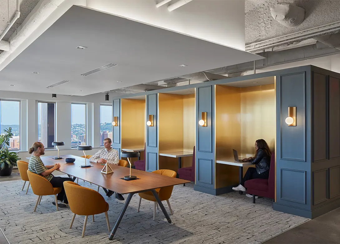 Modern office space with people working at a table and in cozy booths, featuring city views through large windows.