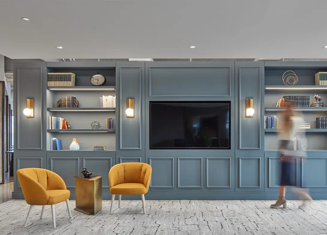Modern office lounge with blue shelves, orange chairs, and a TV. Woman walks by. Stylish, cozy, and inviting workspace.