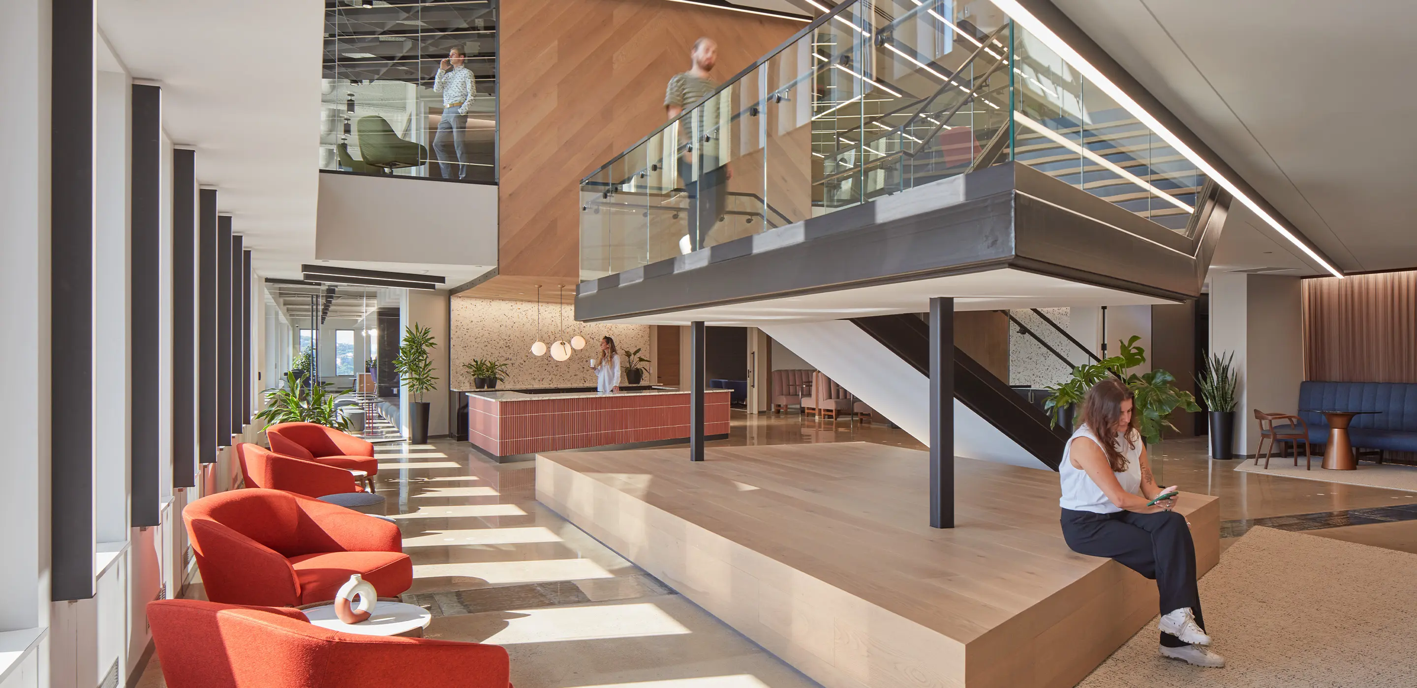 Modern office lobby with red chairs, staircase, and natural light. People working and moving around.