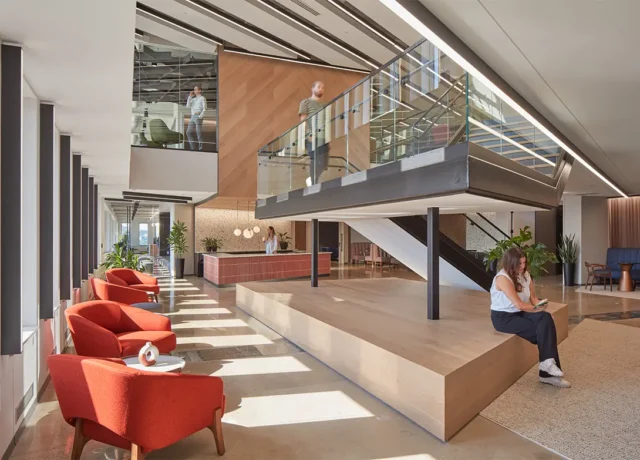Modern office lobby with glass staircase, red chairs, natural light, and plants for a welcoming atmosphere.
