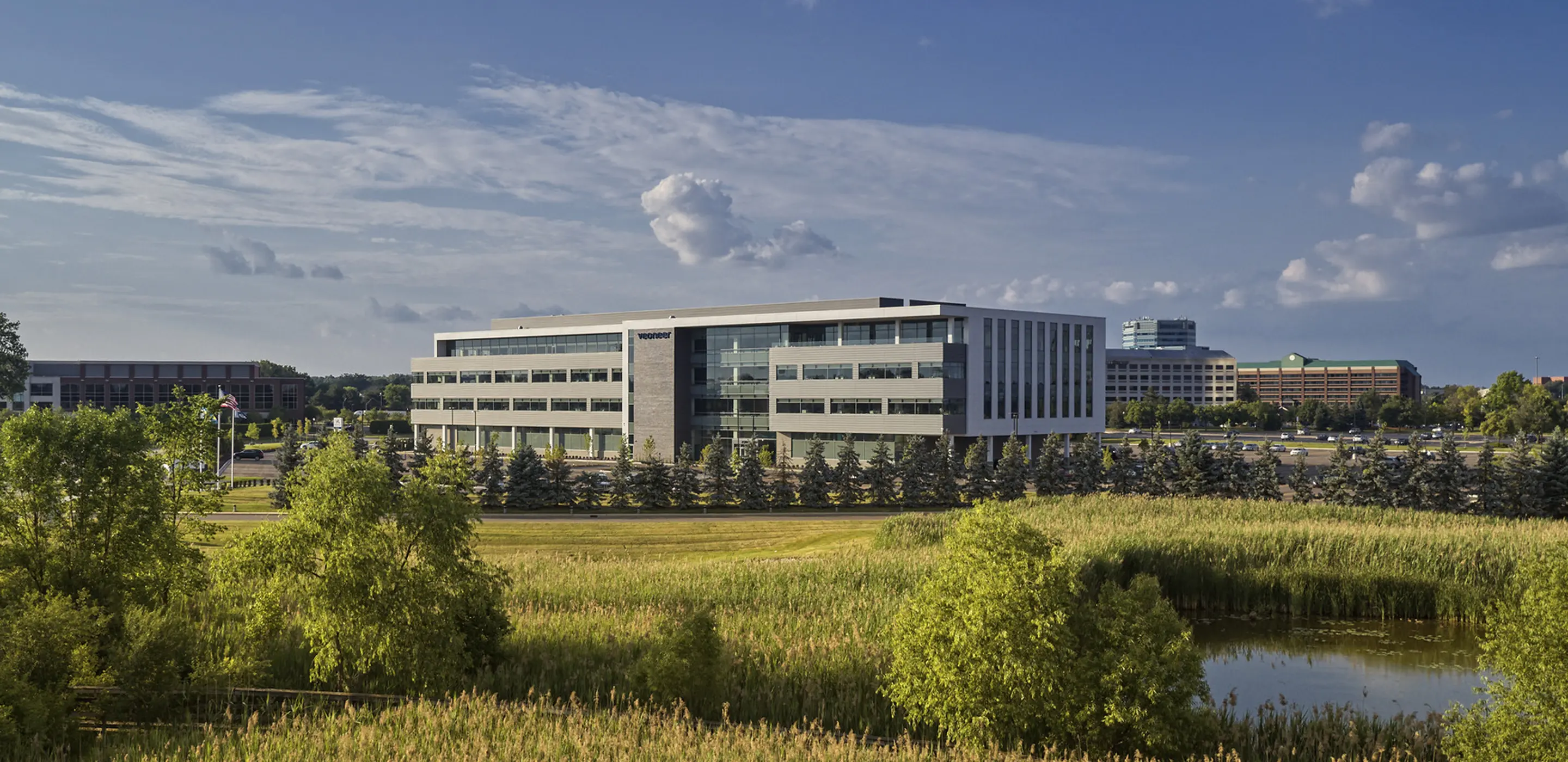 Modern office building surrounded by lush greenery and a pond under a clear blue sky.