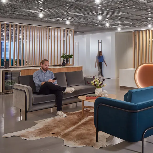 Modern office lounge with a man on a gray sofa and a woman walking, featuring wooden accents and stylish decor.