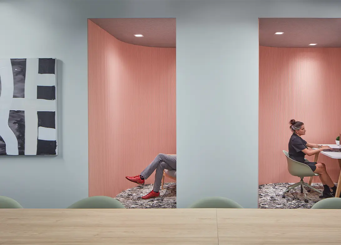 Modern office space with pink alcoves, person working at a desk, another relaxing. Stylish and contemporary design.