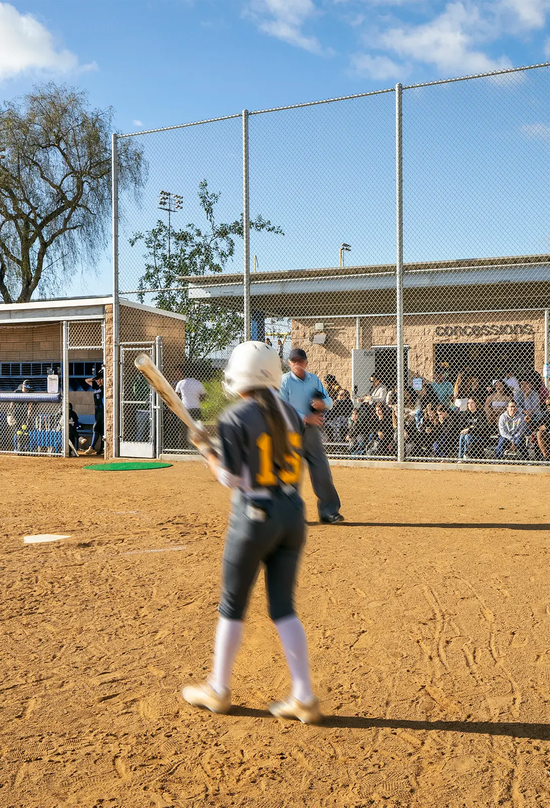 Softball player at bat with helmet, preparing to swing. Umpire and spectators visible in the background at a sunny field.