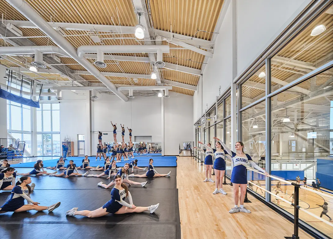 Cheerleading team practicing stunts and stretches in a modern gym facility with high ceilings and large windows.