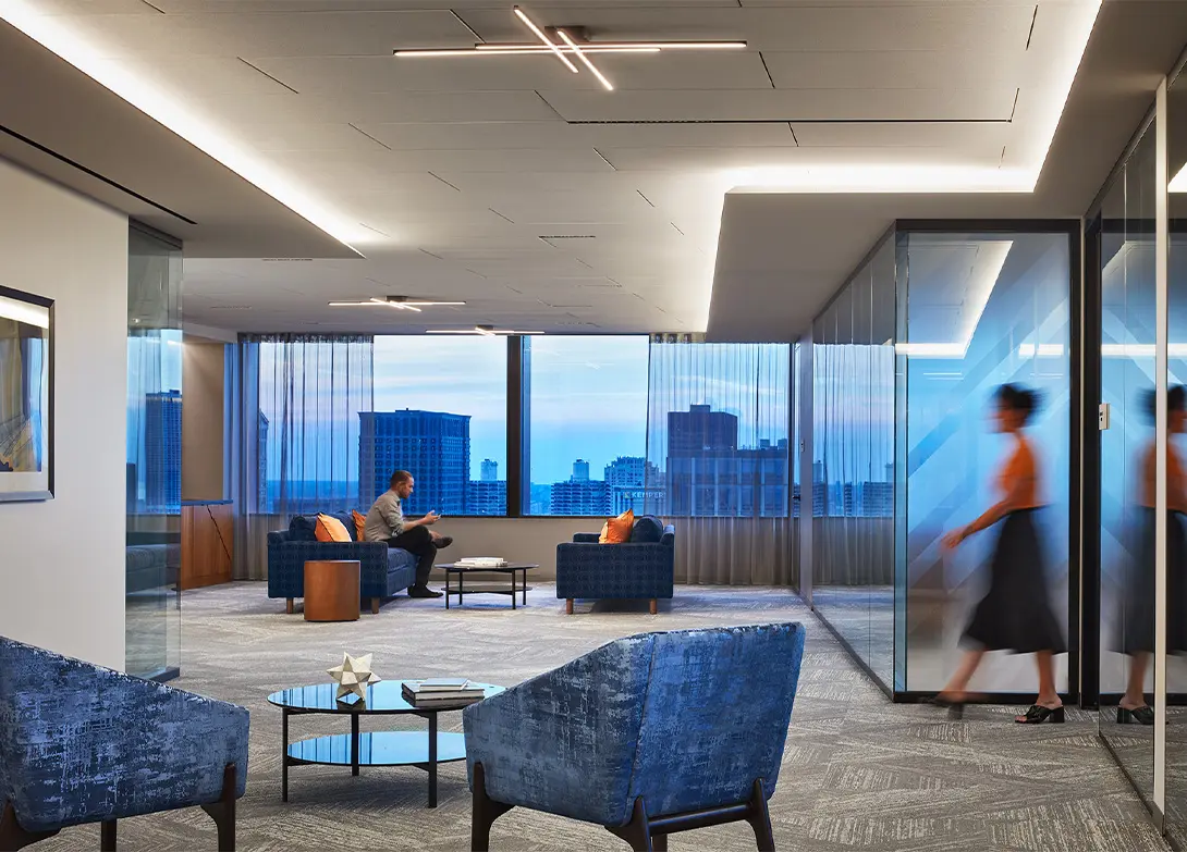 Modern office lounge with a city view, blue chairs, glass walls, and a person walking in the background.