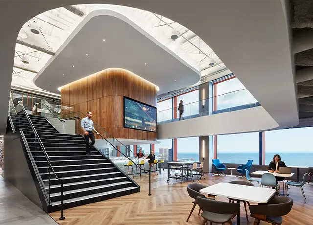 Modern office interior with open space, wooden accents, large screen, staircase, and people working in a bright, airy setting.