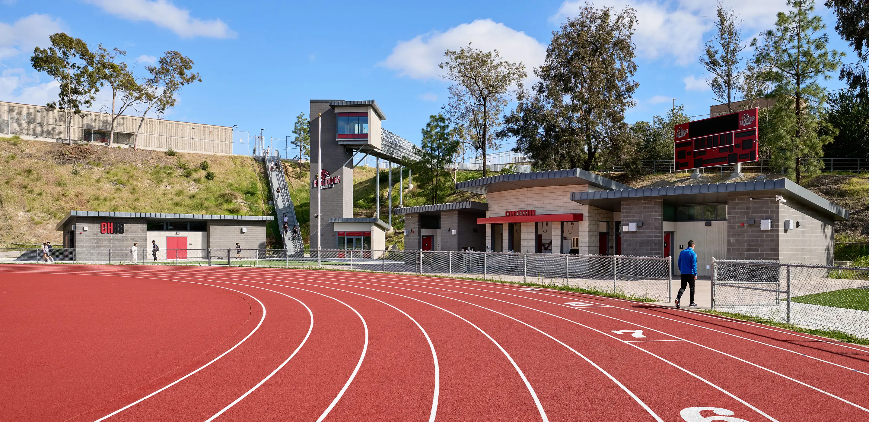 High school track with modern facilities, scoreboard, and walkway on a sunny day.