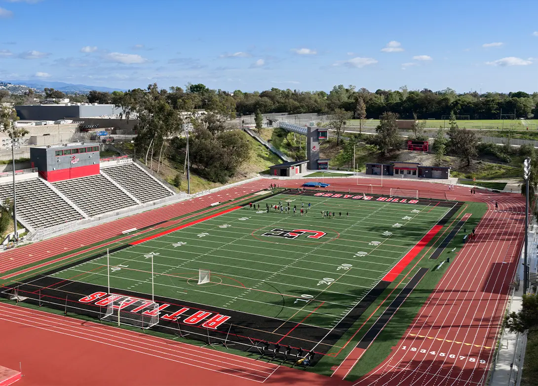 Aerial view of athletic stadium with track and field, surrounded by trees and blue sky, hosting people on the field.