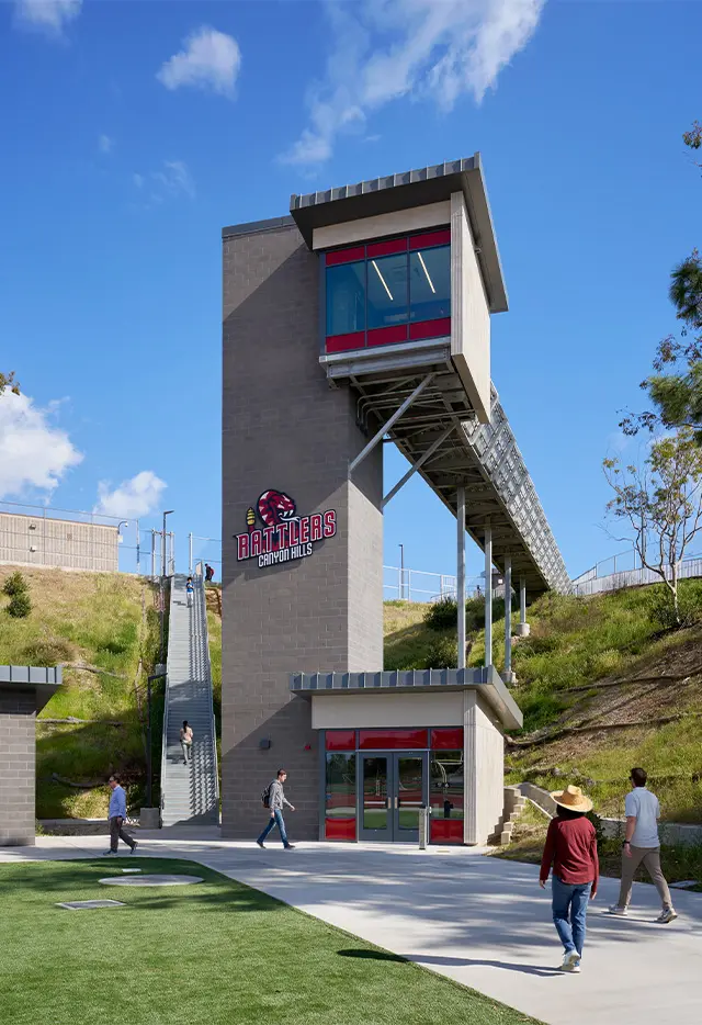 Modern educational building with bridge and stairs, Rattlers Canyon Hills emblem, people walking, sunny day.