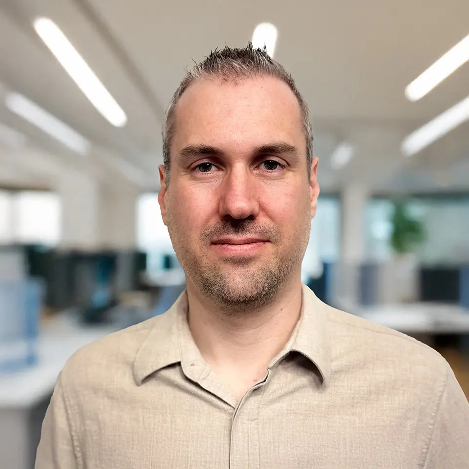 Portrait of a man in a beige shirt smiling in a modern, well-lit office setting.