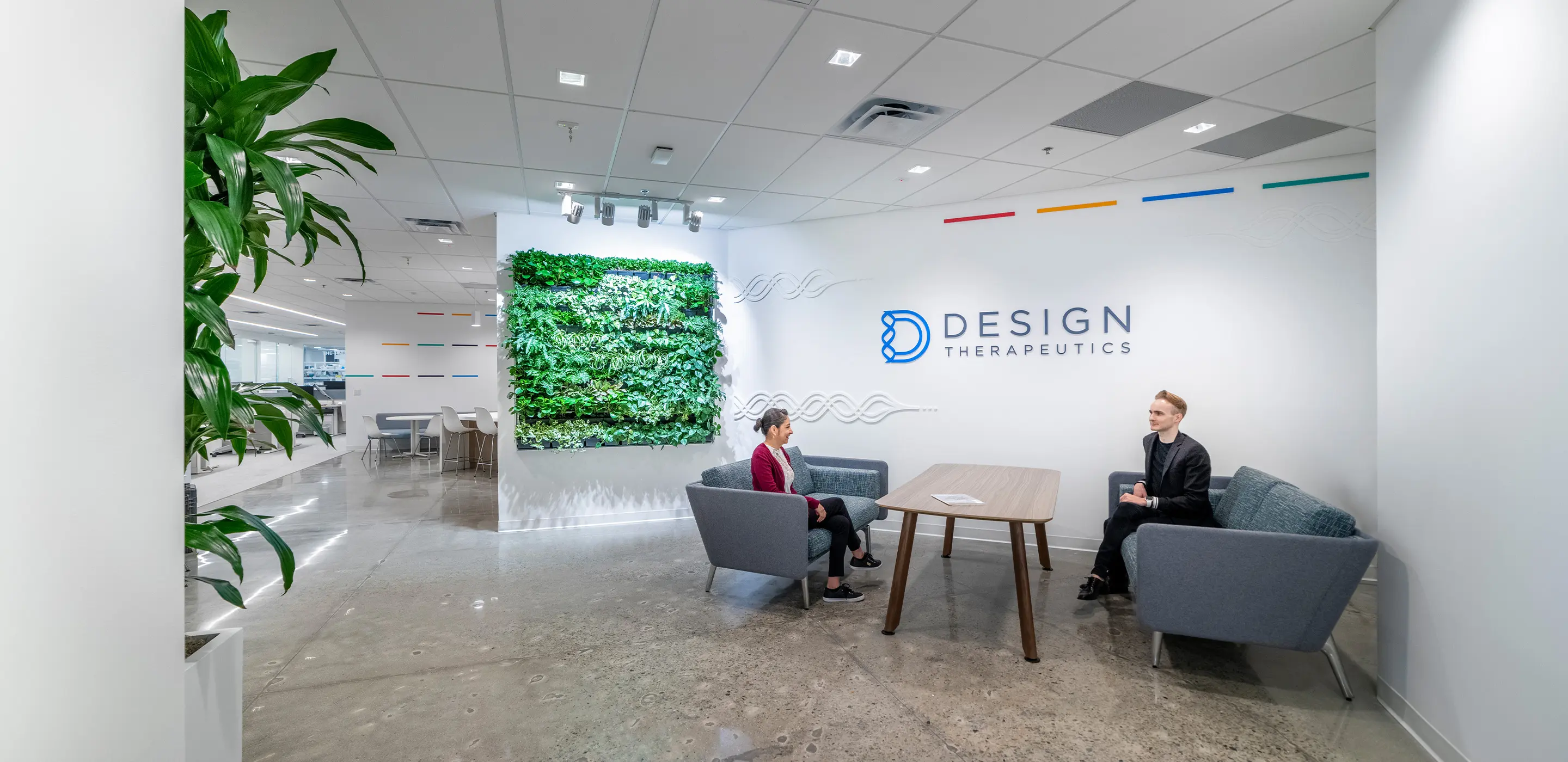 Modern office lobby with green wall, sofa seating, and Design Therapeutics logo. Two people in conversation.