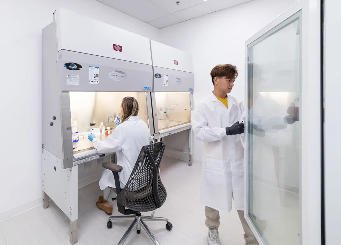 Scientists in a lab, one seated working at a fume hood, another standing nearby in protective gloves and lab coats.