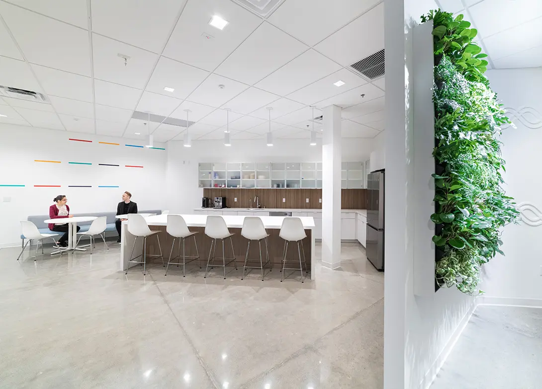 Modern office kitchen with white decor, bar stools, and a green vertical plant wall. Two people are sitting at a table.