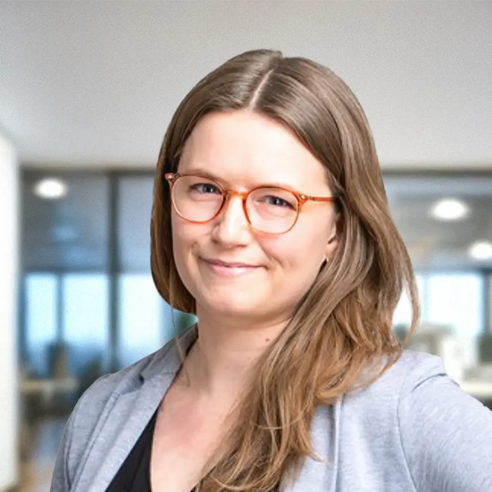 Smiling woman in glasses and blazer in office setting, blurred background view through windows.