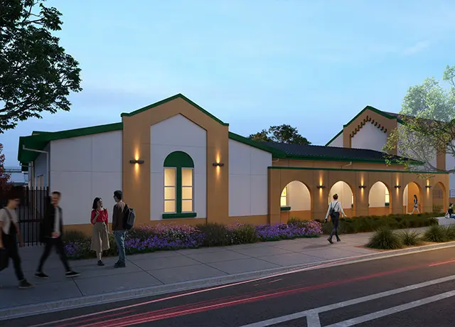 Evening street view of a school building with arched windows, warm lighting, and people walking on the sidewalk.