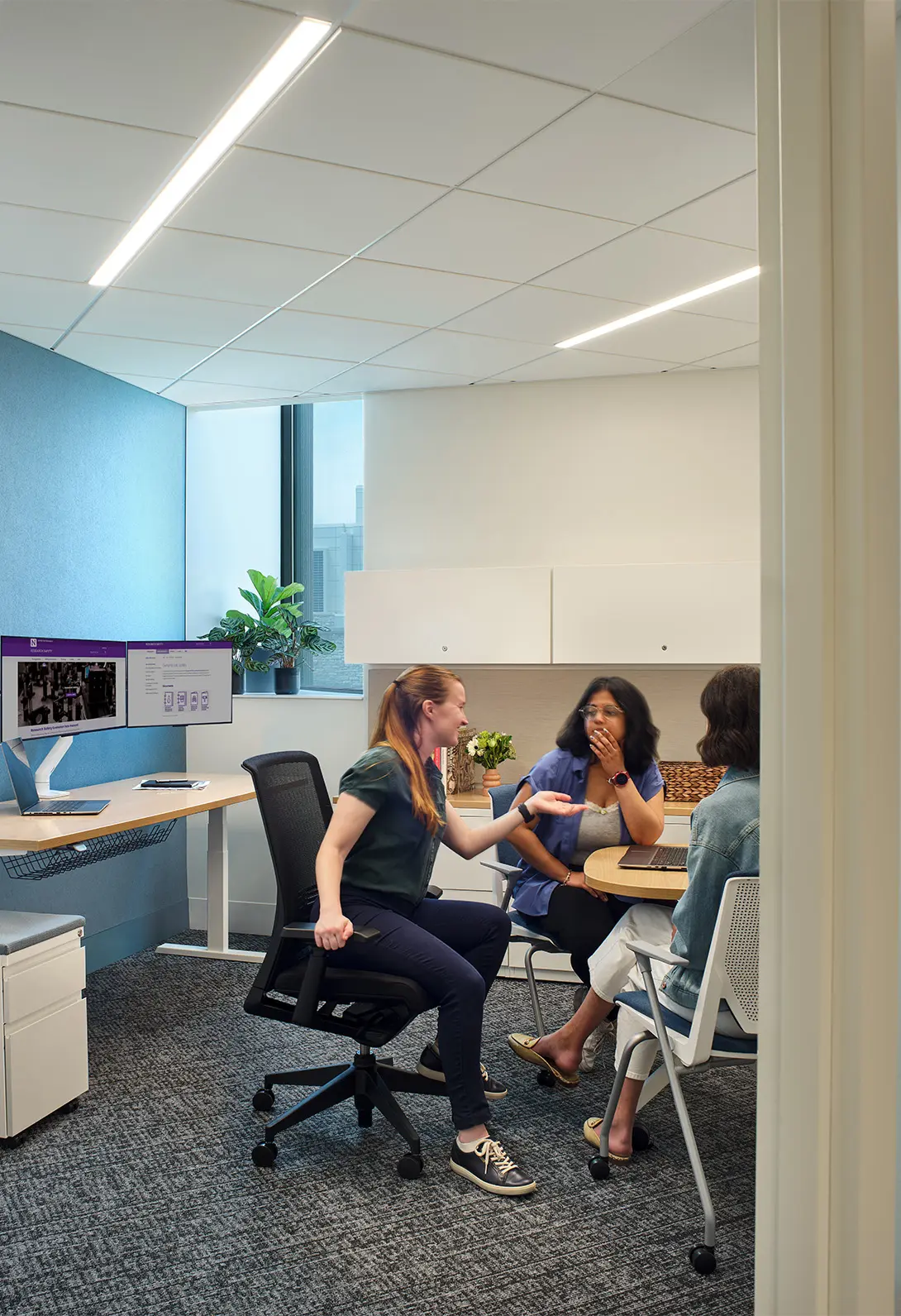 Office meeting with three colleagues discussing ideas in a modern workspace with computers and greenery.