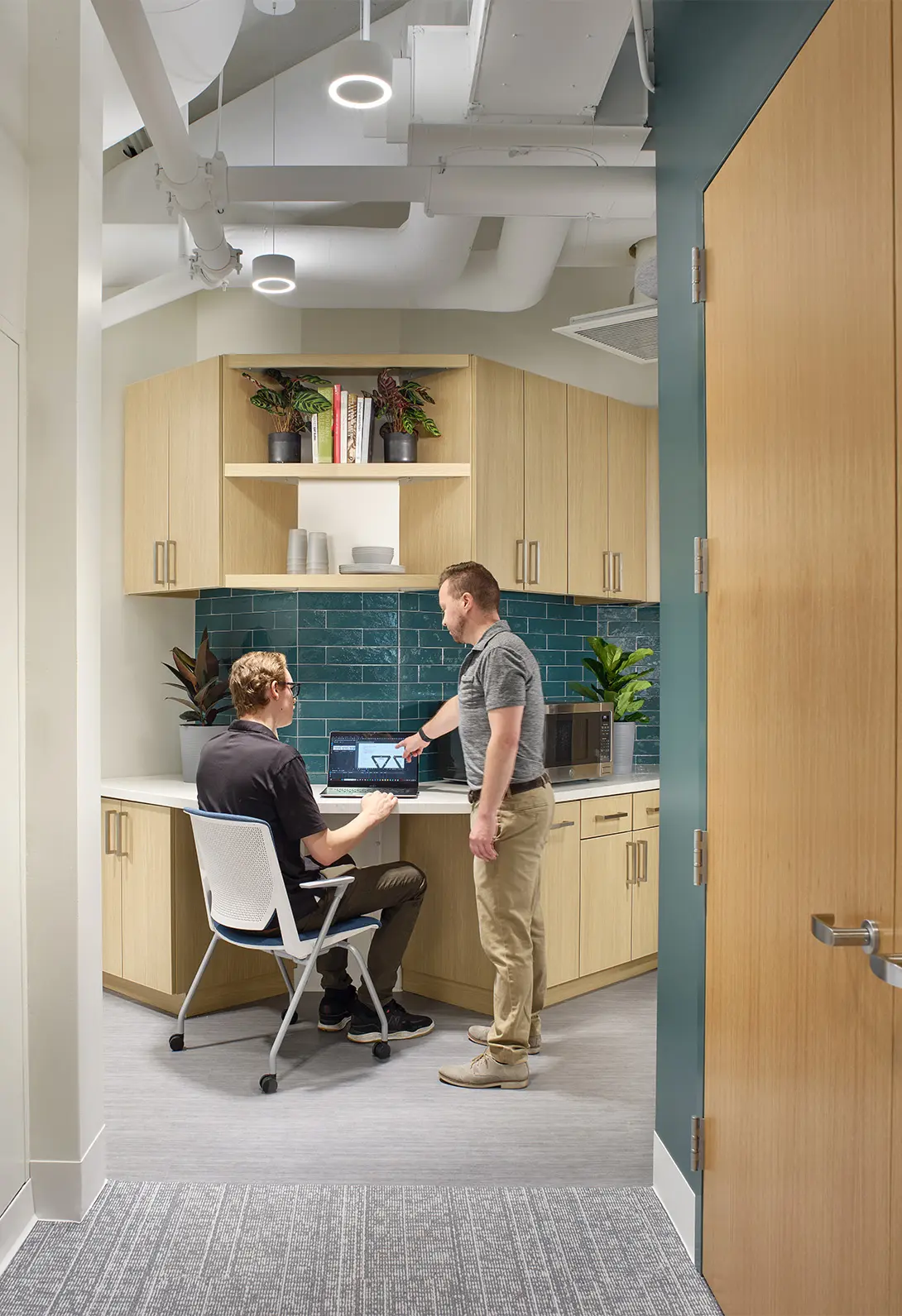 Two people in a modern office kitchen discussing work at a laptop, surrounded by plants and cabinetry.