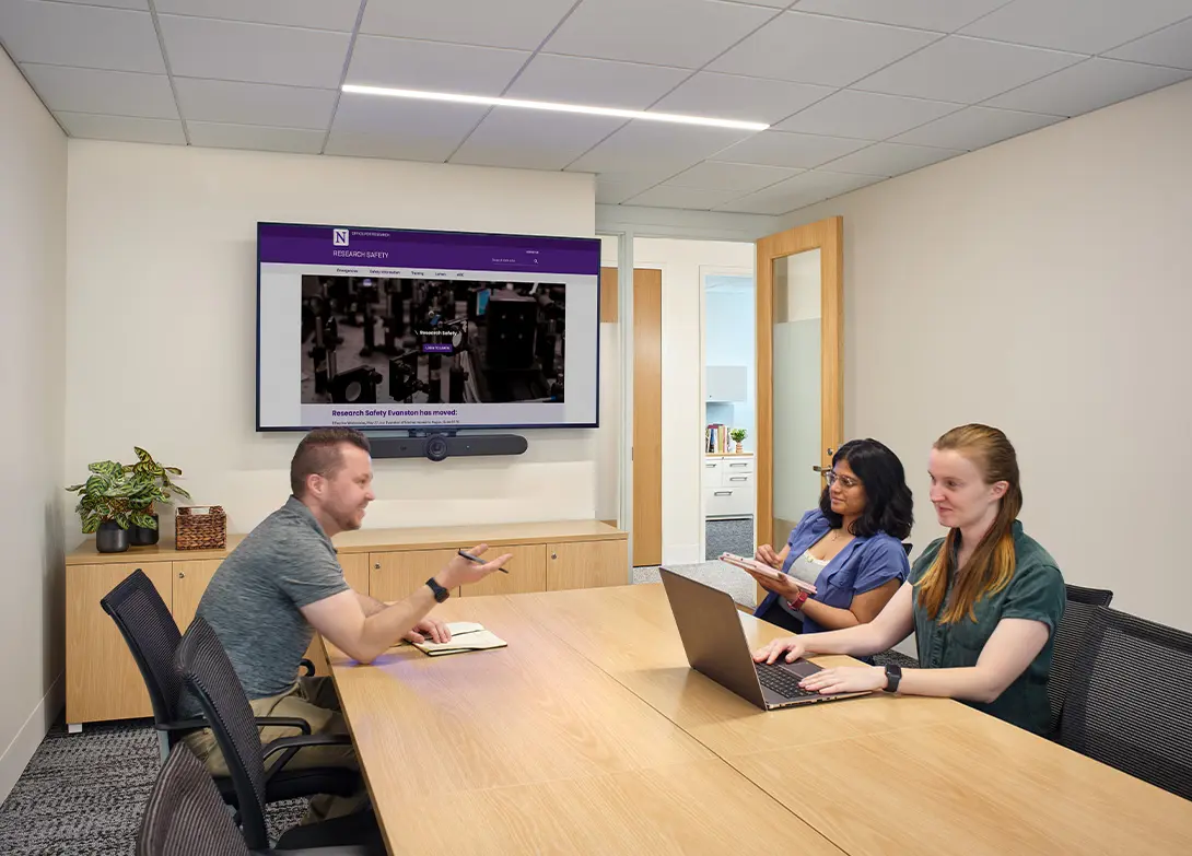 Team meeting in a modern conference room with a large screen displaying a research safety website.