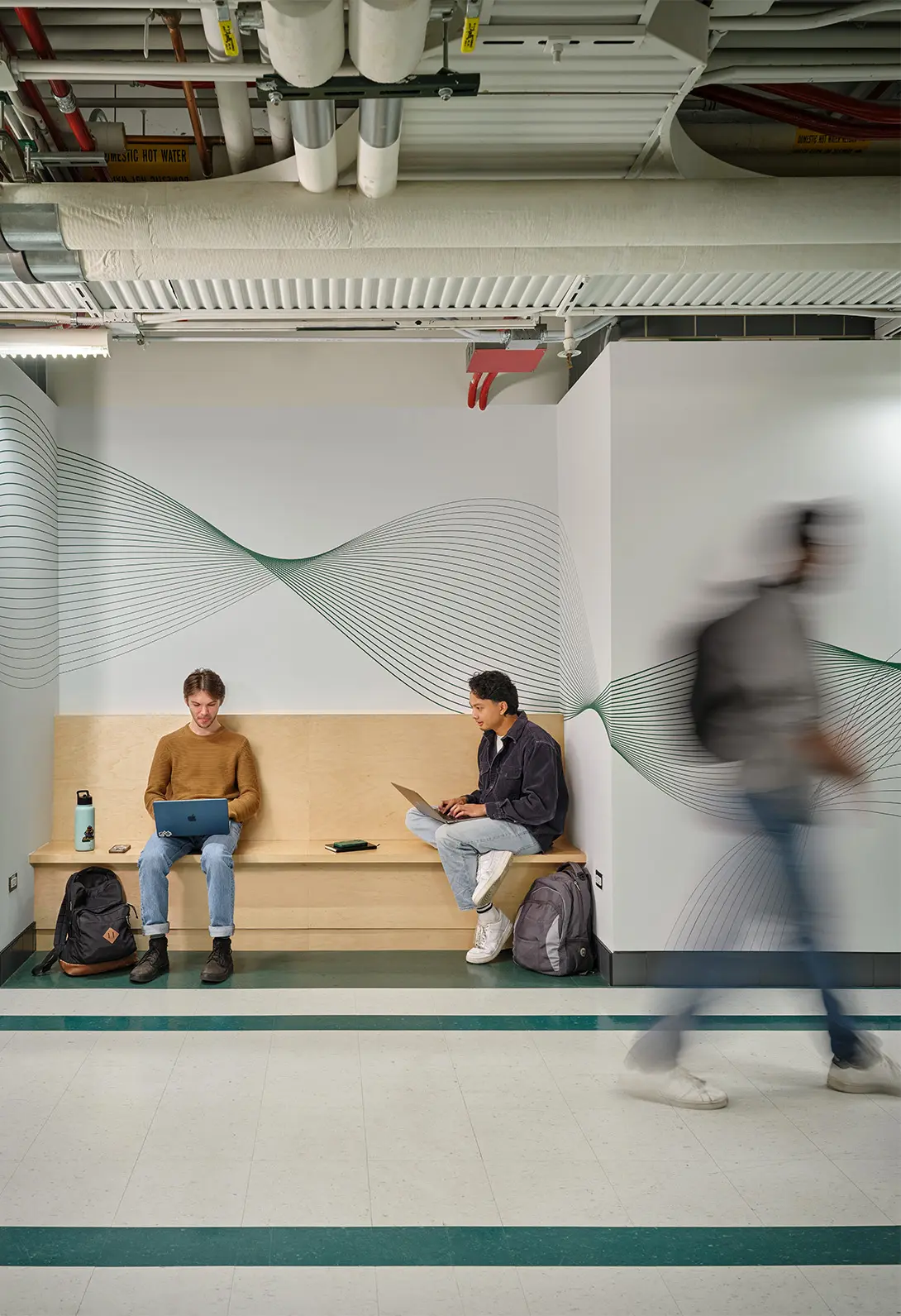 Students sitting on a bench with laptops in a modern hallway, abstract wall art, motion blur of a passerby.