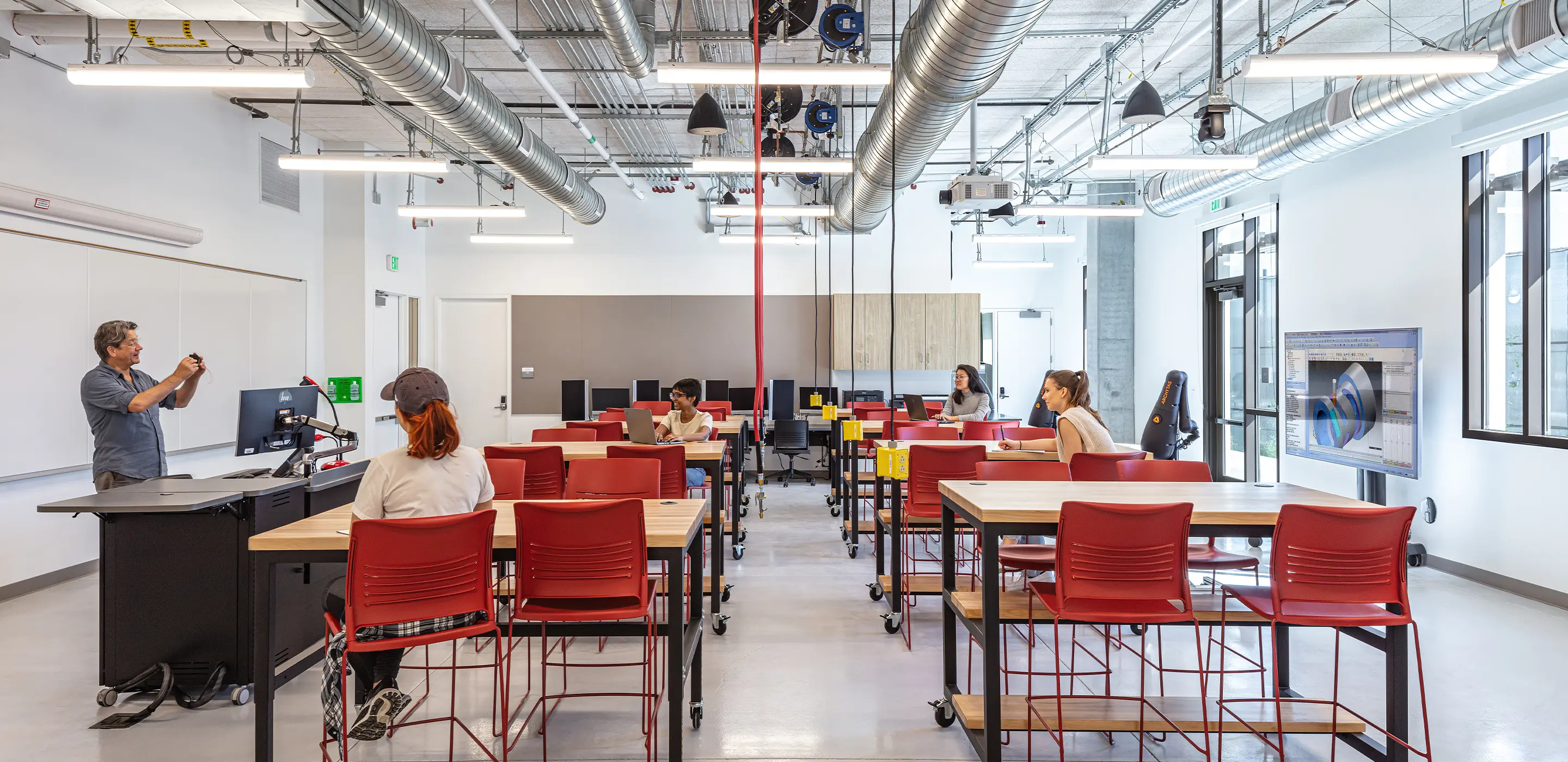 Modern classroom with students at desks, teacher presenting, red chairs, and computers with a projection screen.