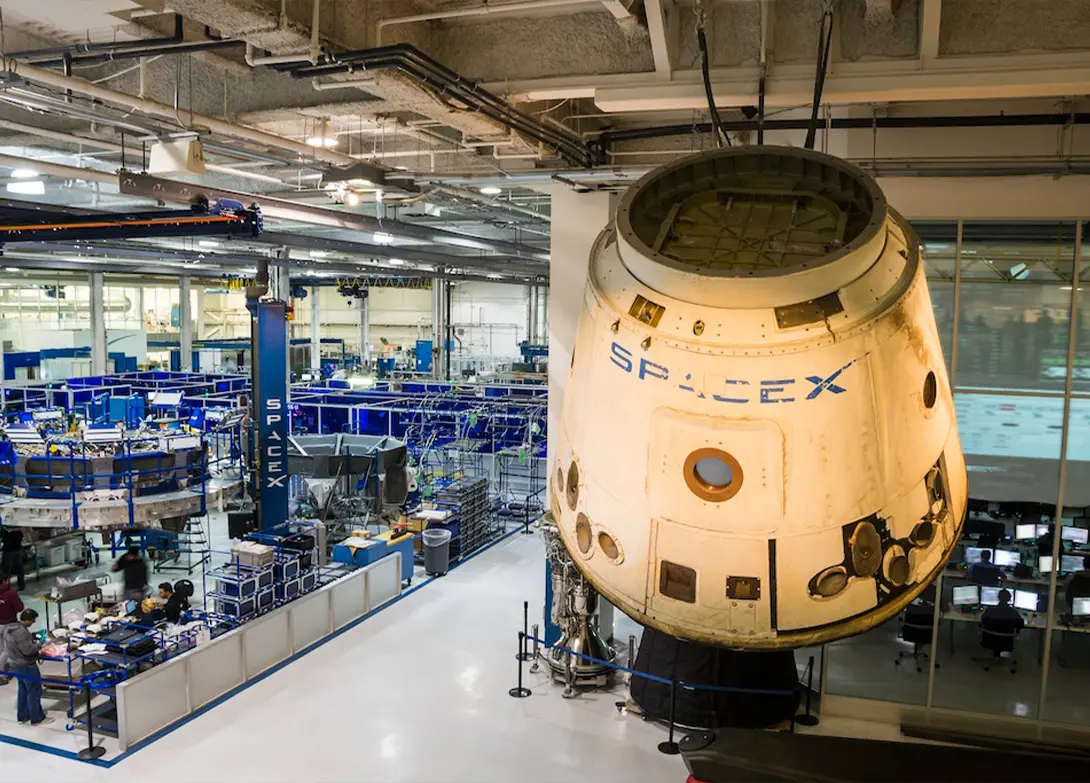 SpaceX spacecraft displayed above a busy, high-tech manufacturing facility with workers and machinery.