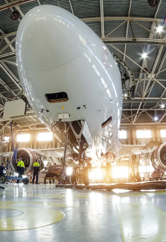 Commercial airplane nose in hangar, undergoing maintenance with workers, engines, and sunlit backdrop.