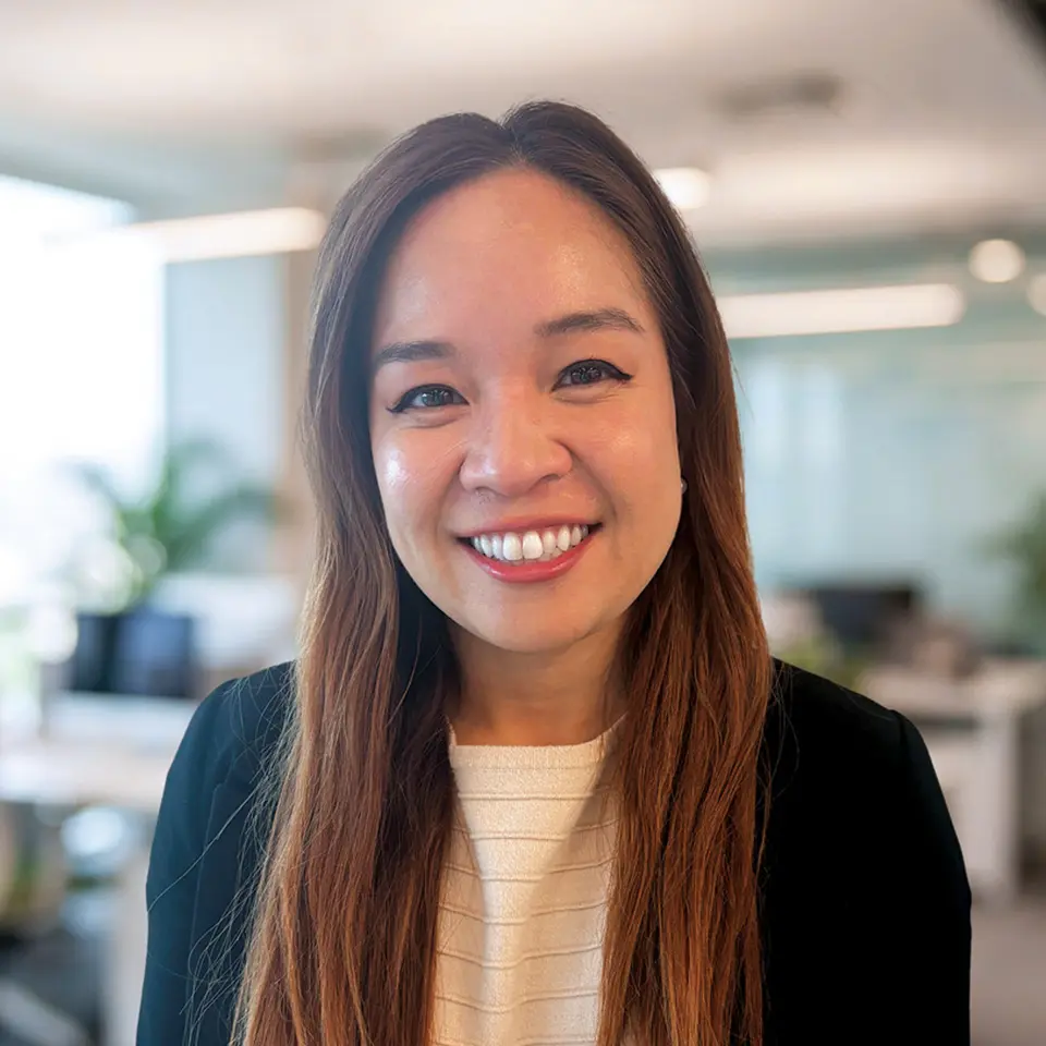 Smiling professional woman in a modern office setting, long hair, wearing a blazer and white top.