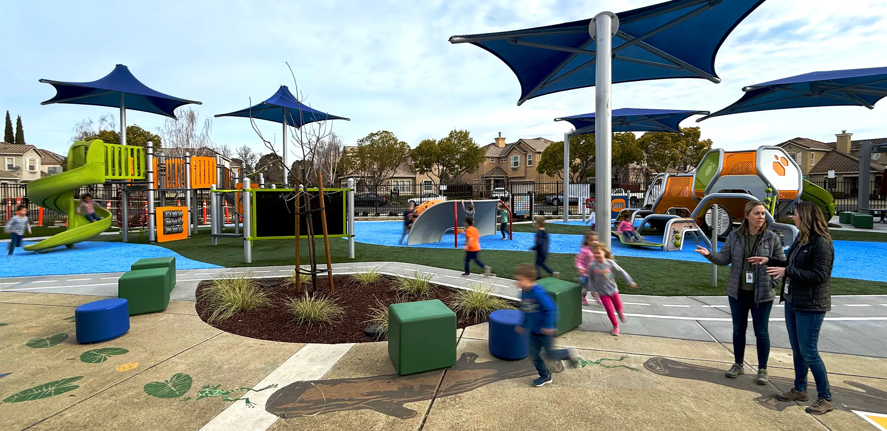 Modern playground with colorful equipment, children playing, and adults talking under blue shade structures. Urban setting.