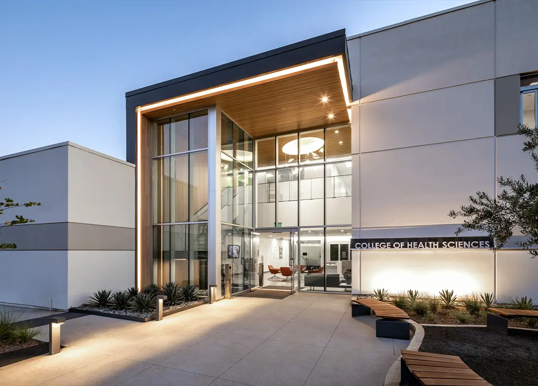 Modern College of Health Sciences building entrance with large windows and evening lighting.