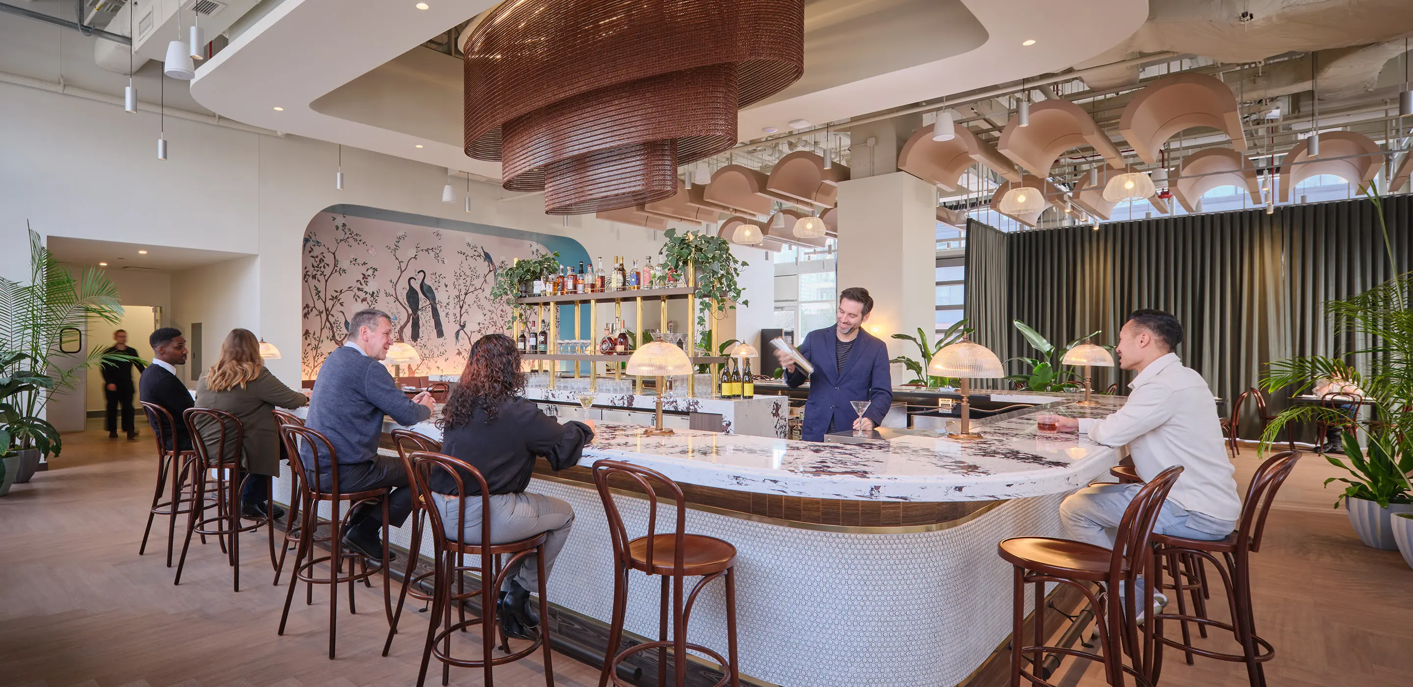 Modern bar with people seated and bartender mixing drinks, featuring elegant decor and plants.