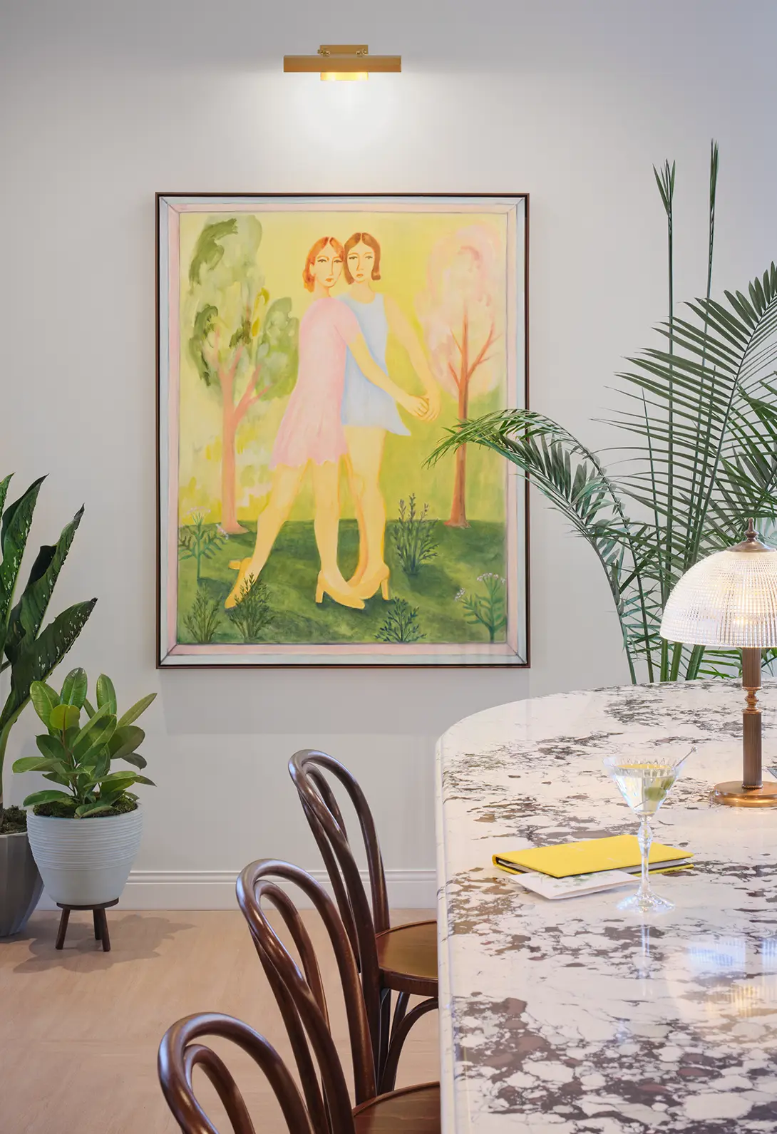 Framed art of two figures with plants and marble countertop in a stylish room, featuring warm lighting and wooden chairs.